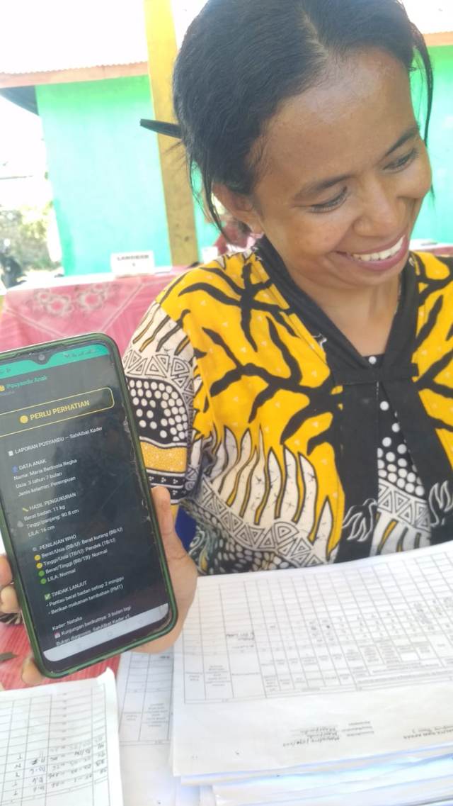 A kader at a Posyandu table smiles while showing a child's result on her phone